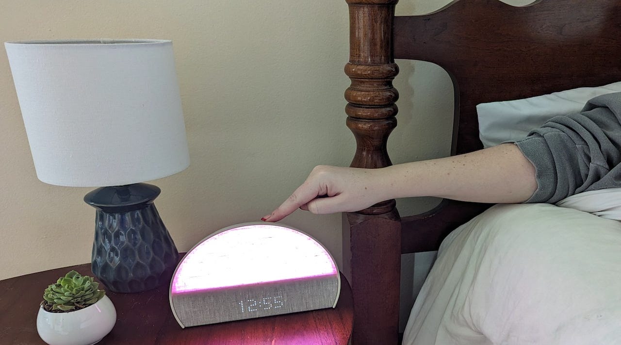 Person in bed pushing a button on a lit-up Hatch Restore 2 device on a nightstand