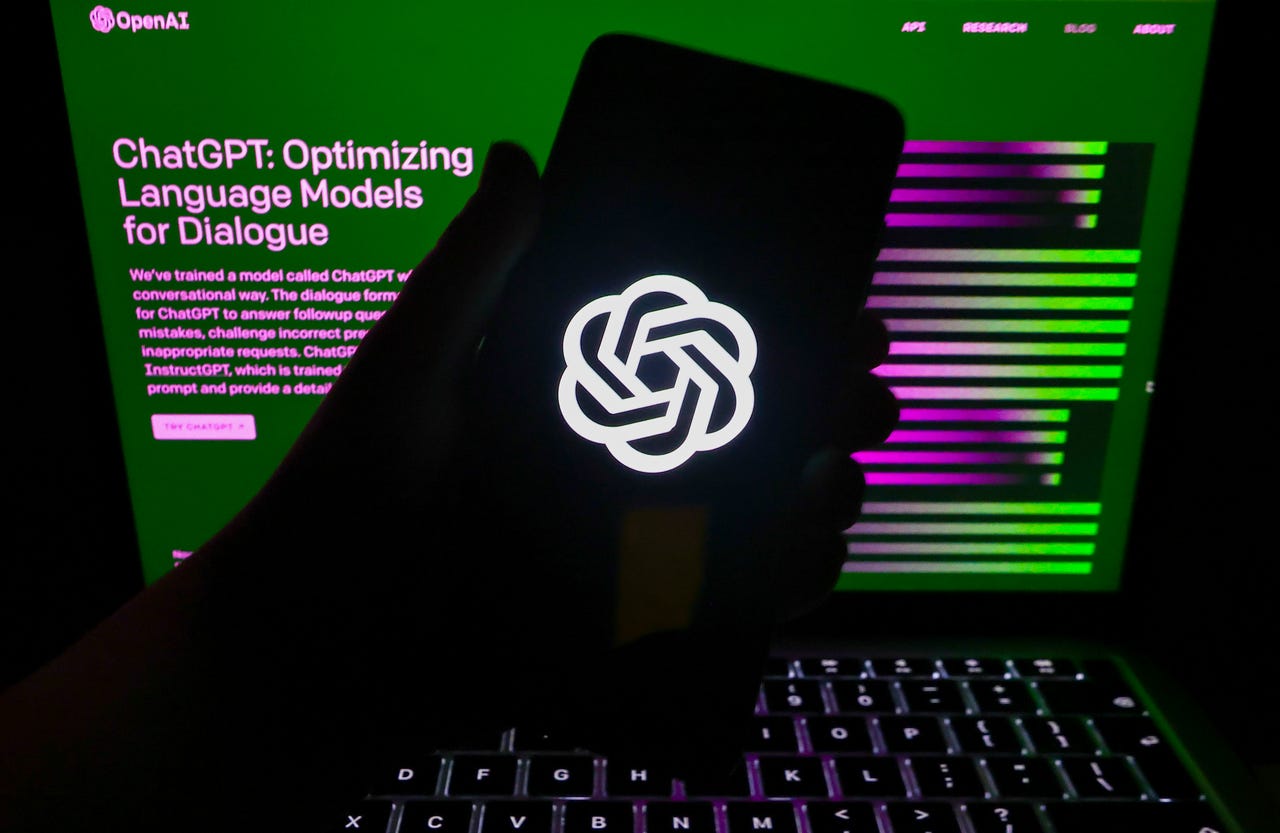Silhouette of hand holding phone with OpenAI logo in front of laptop screen showing ChatGPT