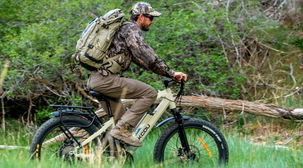 A man in camouflage and wearing a hiking backpack on a Bakcou eBike, pedaling through a woodland clearing